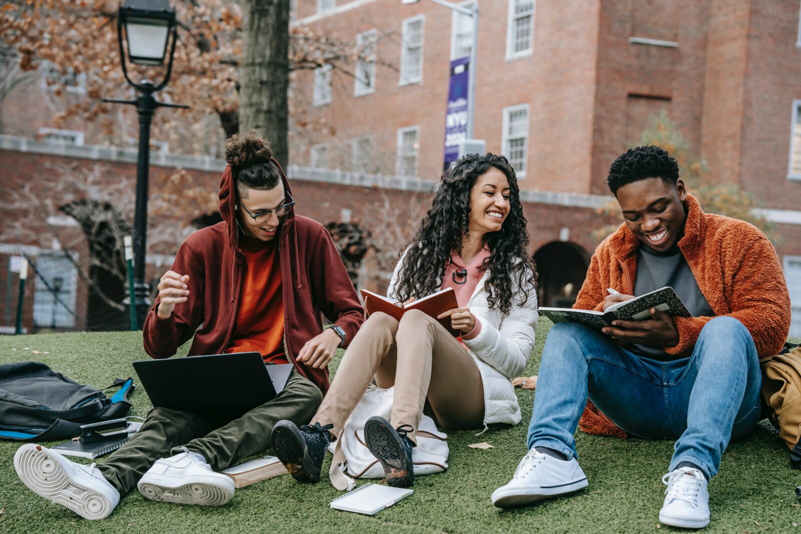 A group of three young people siting on the grass smiling laughing and studying.